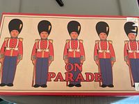 Box lid showing the game's title 'On Parade' and illustrations of five cardboard soldiers in red coats and tall black helmets.
