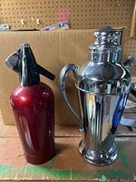 Red soda spritzer bottle and chrome cocktail shaker set on wooden surface