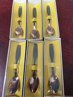 Photo of six memorial spoons individually boxed with yellow interior and clear plastic cover, spoons have metal bowls and dark handles featuring designs related to Lyndon B Johnson.