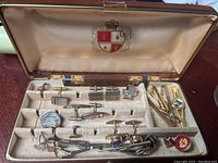 Open box view showing over 20 pieces of men's jewelry including cuff links, tie pins, and pins organized in a velvet-lined storage box with emblem on the lid.