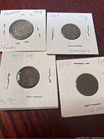 Photo showing four old European coins in white cardboard holders placed on a wooden surface, labels handwritten with year and denomination visible.