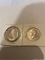 Two 1976 US Kennedy half dollar coins individually encased in white cardboard holders, one showing Independence Hall on the reverse and the other showing Kennedy portrait on the obverse.