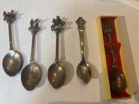 Five souvenir collectible spoons on white background, including boxed Oregon spoon and Nashville musician figurine spoons showing detailed top designs and engraved bowls.