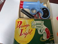 Top view of the blue and red plastic toy pistol inside original box with black accessory and visible Morse code instructions underneath.