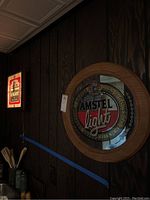Dimly lit photo showing Amstel Light circular mirror plaque on dark wood panel wall with wooden frame and reflective center showing logo text and colors. Budweiser Light illuminated clock is visible in the background.