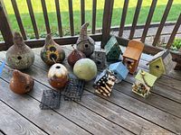 Photo of all seven gourd birdhouses (with various painted designs) and seven wooden/resin/tin birdhouses (various colors and styles) along with two black wire suet feeders on a wooden deck.