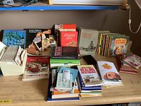 Overview of assortments of cookbooks, children's books, religious books, and recipe cards arranged on table showing wear.