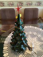 Full view of the ceramic Christmas tree on a glass table with crochet tablecloth and chairs in background.