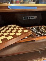 Wooden chessboard partially open showing the board surface and edge of the wooden case with chess pieces inside.