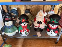 Seven Christmas-themed sculptures arranged on a metal shelf in a wood-paneled room. Includes three resin figures and four hand-painted ceramic snowmen. Colors include red, black, green, white. Tallest piece around 16 inches.
