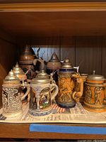 Nine lidded ceramic beer steins arranged on a shelf showing various designs and colors including blue, brown and cream with relief decoration and pewter lids