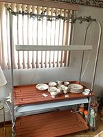 Full view of metal frame growing station with brown plastic trays holding some dishware (not included), overhead light fixture, and window blinds in background.