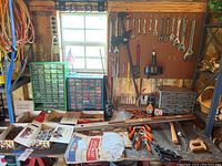 Overall view showing three hardware organizers with many drawers, pegboard above with various wrenches, clamps, level, and other tools and parts on table and shelves