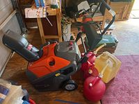 Full view of snow blower unit, three red gas cans, one yellow gas can and a funnel on a shop floor.