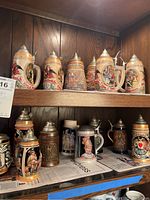 Full collection of ceramic beer steins displayed on wooden shelves, some with metal lids, showing variety of designs and colors.