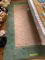Full view of the rug next to a bed showing the beige center and green border, with visible wear and pet-friendly home signs.