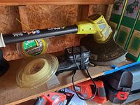 Photo of Ryobi weed trimmer showing battery insert area, trimmer head, two batteries, charger, and spools of line on shelf.