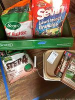 Photo showing unopened Scotts Turf Builder, Sevin Lawn Insect Granules, Scotts Step 4 Fertilizer, green metal spreader, partial grass seed container, and books.