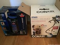 Photo of Bissell SpotClean ProHeat spot cleaner in box along with Conair Ultimate Fabric Steamer box on floor.