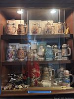 Full view of glass cabinet shelf showing ceramic steins on top two shelves and decanters on bottom shelf