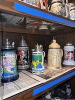Six ornamental beer steins displayed on a shelf with attached pewter lids. Designs include maps and traditional scenes.