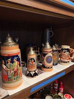 Photo showing eight beer steins and mugs on a shelf, several with ornate pewter lids and detailed painted scenes representing Bavarian folk themes and historical motifs.