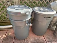 Three gray Rubbermaid trash cans with lids side by side on outdoor pavement with lattice background. Cans show light dirt and scratches.