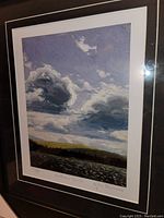 Framed fine art print showing scenic clouds over a body of water and landscape with visible signature and title on the print border.