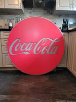 Front view of the round Coca-Cola promotional display sign against kitchen cabinets, showing the vibrant red and white logo.