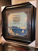 Framed silkscreen print showing a waterfront with boats and a steamship, featuring text advertising Dominion Express Travellers Cheques.