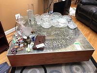 Square coffee table with dark wood laminate base and metal edges. Top features detailed Medieval-themed bas relief in circular design. Various glassware, bowls, and decorative items displayed on top, not included.