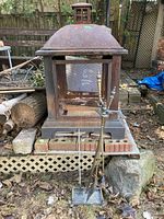 Front view of the rusty metal fire pit with mesh sides, set on bricks outdoors, with fireplace tools in front.