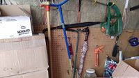 Photo showing various garden tools hanging on a pegboard wall including a manual tiller, weeder, caulking applicator, brush and suction cups, plus a green garden hose nozzle and blue handled tools.