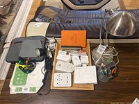 Lot of office supplies on small wooden tray and surrounding area including stainless steel desk lamp, power strip, carbon monoxide alarm, pens, stapler, cables, and paper.