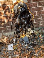Bundle of outdoor string lights coiled and resting against a brick wall among fallen dry leaves, showing multiple bulbs and cables tangled together.