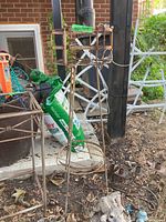 Photo of a tall rusty metal plant stand with rounded and square design elements, a green plastic pot filled with dirt, and bags of turf builder fertilizer in an outdoor garden setting.