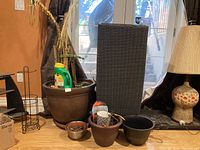 Photo showing multiple plant pots including a large brown ceramic pot with soil and dead plant, various smaller pots, a wire plant stand, and a lamp in background.