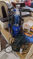 Full view of blue Kobalt power washer unit with wrapped hose and spray gun placed on floor next to a chair.