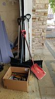 Photo showing four shovels, one metal and three plastic, with wood handles, leaning against wall next to a box containing accessories.