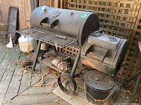 Front and side view of Oklahoma Joe's offset smokey grill with side firebox, metal bucket below and aroma chips on shelf. Outdoor setting on wooden deck.