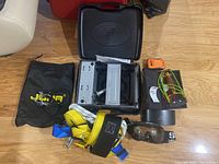Photo showing ski goggles, emergency beacon, slingshot, camping stove case, and JOHA safety harness with rope and carrying bag on wooden floor.