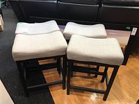 Four metal frame stools with light beige upholstered seats arranged together on floor showing overall condition and shape.