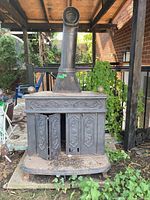 Full frontal view of weathered metal wood stove positioned outdoors with chimney pipe and ornate front doors partially open