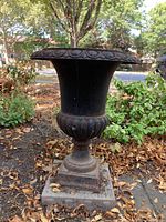 Full view of the weathered dark cast iron urn planter with decorative scalloped rim and base, surrounded by fallen leaves and garden plants.