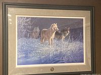 Full view of the framed print showing three wolves in a snowy setting with frosted plants. The frame is black with double matting in gray and cream.