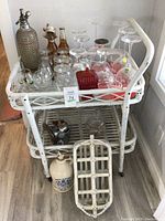 Full view of white wicker tea cart with wheels holding multiple glassware items, seltzer bottle, metal corkscrew, red glass candy dish with lid, and white wicker beverage tray leaning at front.