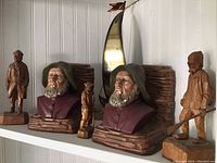 Shelf display with three carved wood figures, two painted ceramic bookends depicting older men in coats, and a metal sailboat figurine