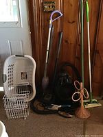 Lot of cleaning supplies and appliances aggregated against a wood wall, showing Shark vacuum, floor mops, laundry basket, and wire baskets.