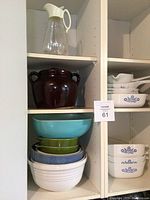 Shelf with large turquoise, green, blue ribbed, and white ridged mixing bowls; brown pottery bean pot with lid and large glass pitcher with white plastic spout and handle.