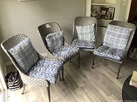 Four wicker style dining chairs with black metal legs and removable seat and back cushions in blue and white patterns, arranged around a corner in a room with grey wood flooring.
